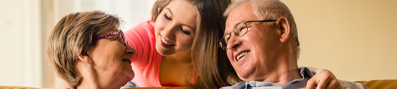 Two senior citizens sitting on a couch. Behind the couch is a younger girl who may be a relative.