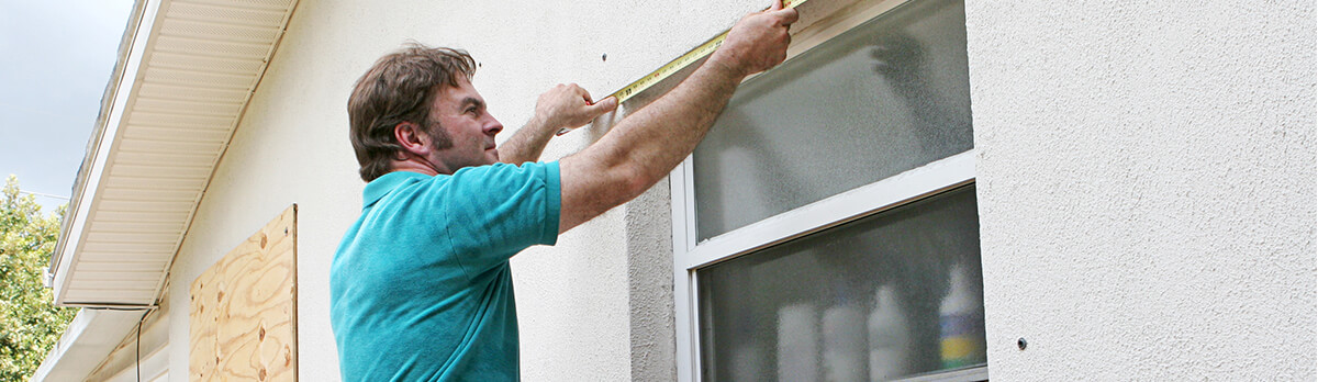 Man on a ladder, outside a home, measuring the top of a window so he can attach plywood to prepare for a hurricane.