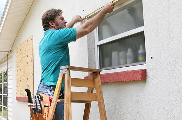 Man on a ladder, outside a home, measuring the top of a window so he can attach plywood to prepare for a hurricane.