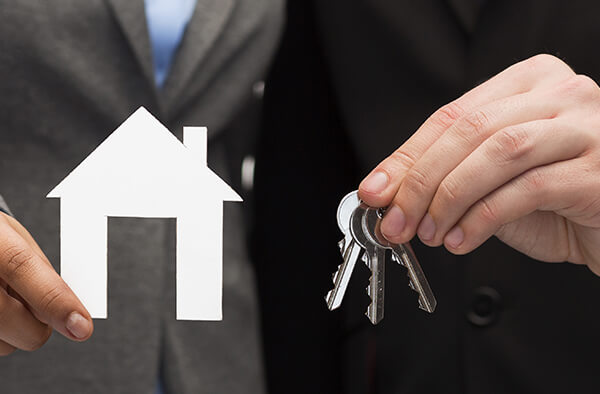 Man holding a set of house keys in one hand and a small paper house cut out of paper in the other hand.